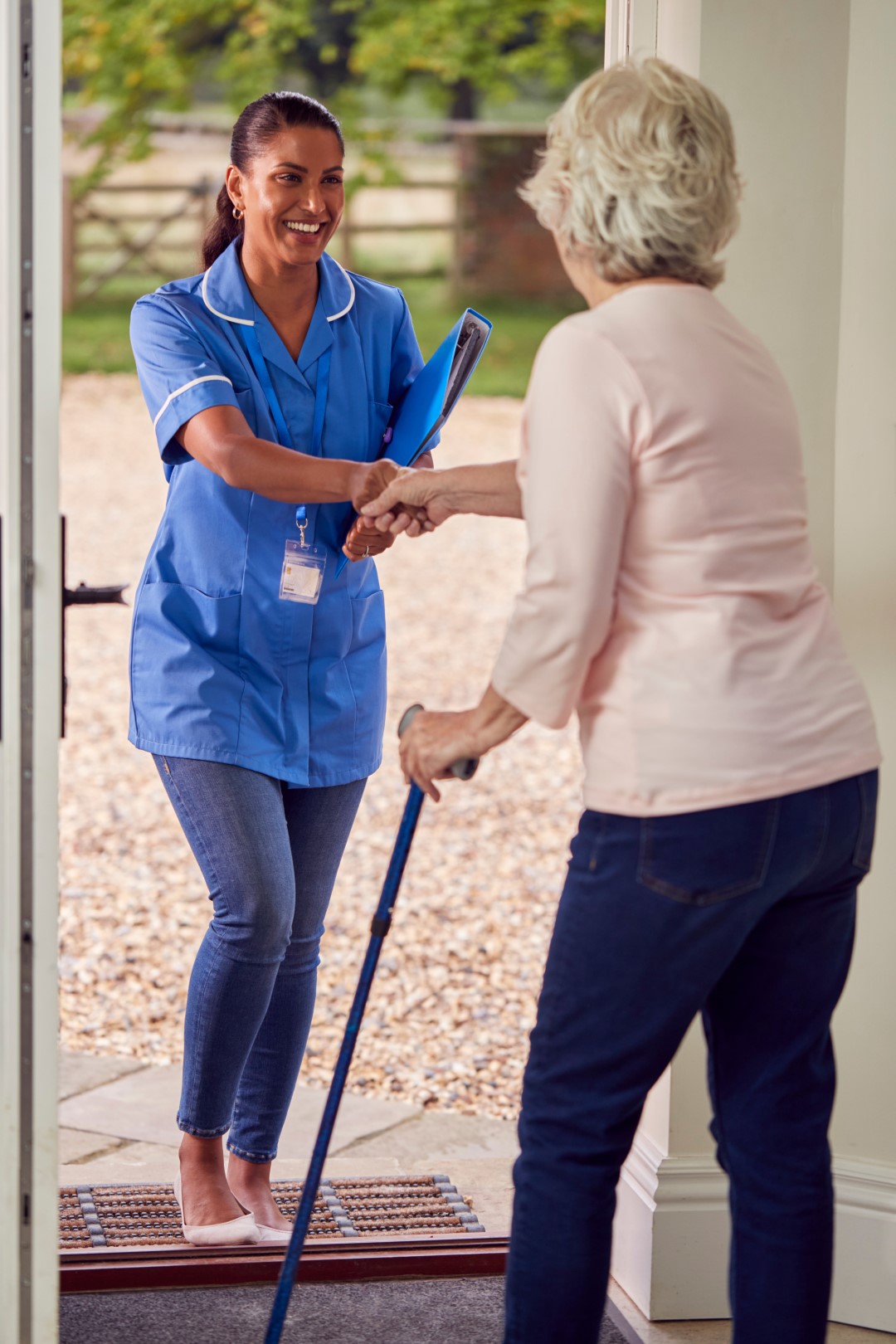 Senior Woman At Home Using Walking Stick Greeting Female Nurse Or Care Worker In Uniform At Door