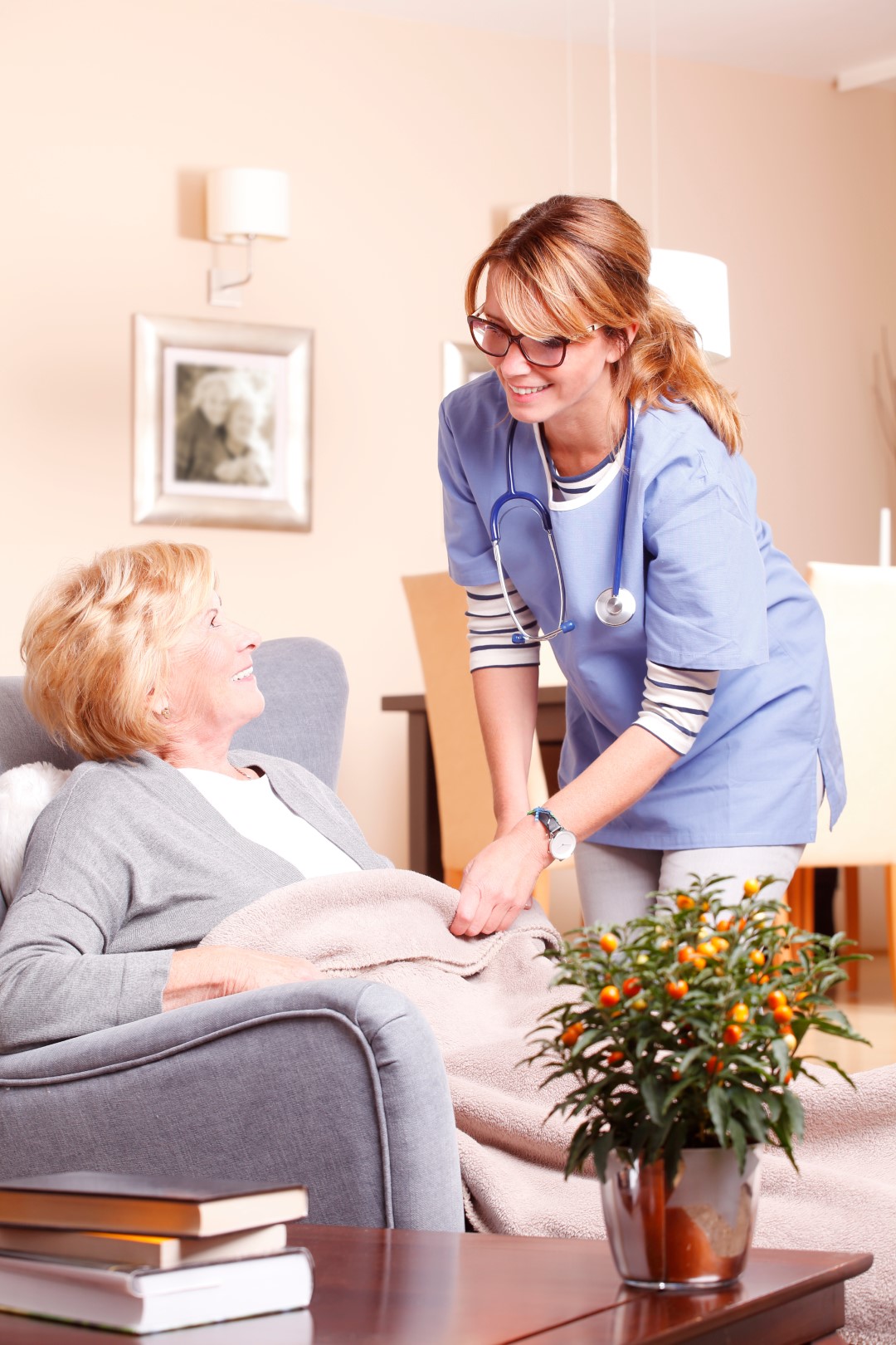 Portrait of old woman sitting at home in armchair while smiling caregiver covering her with blanket